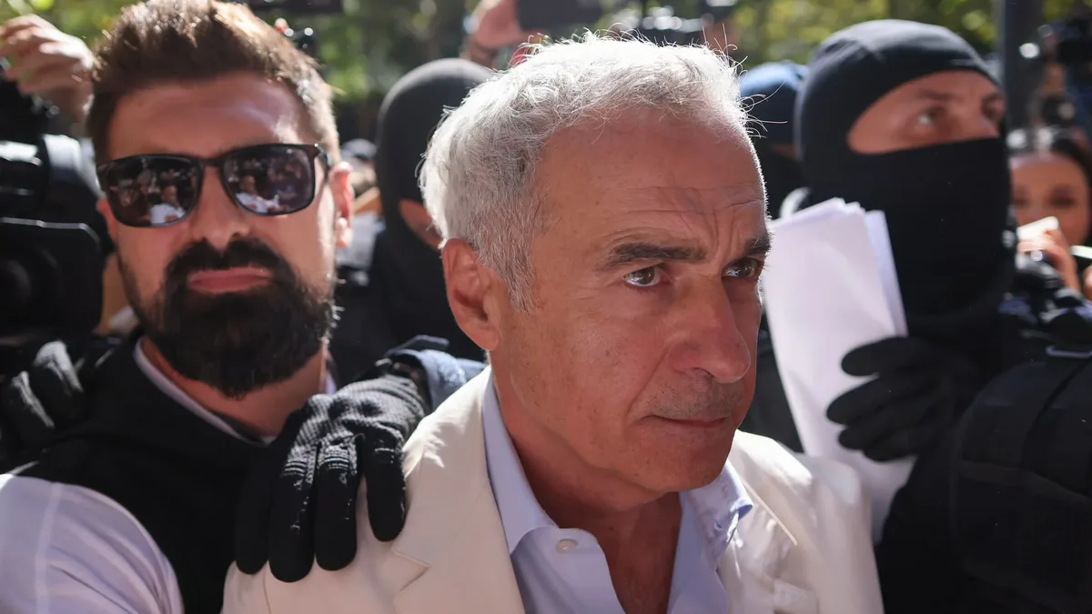 Călin Georgescu, în sacou deschis, înconjurat de persoane cu echipament tactic și camere de presă, în aer liber.
