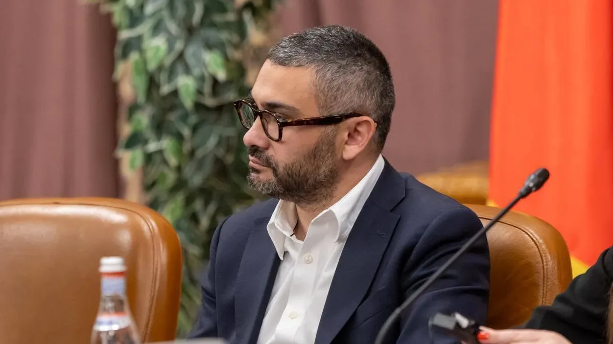 Florin Manole, în costum bleumarin și cămașă albă, așezat la o masă de ședință, lângă un microfon.