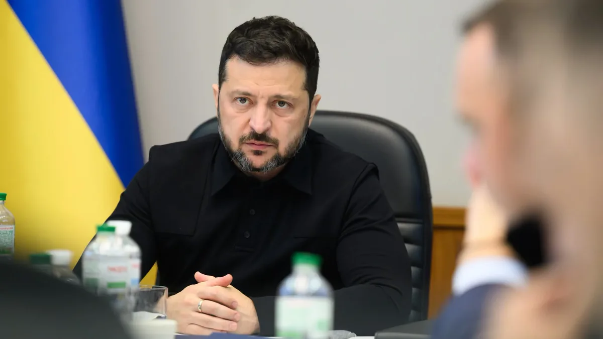 Volodîmîr Zelenski la o masă de reuniune, cu mâinile împreunate și drapelul Ucrainei în fundal.