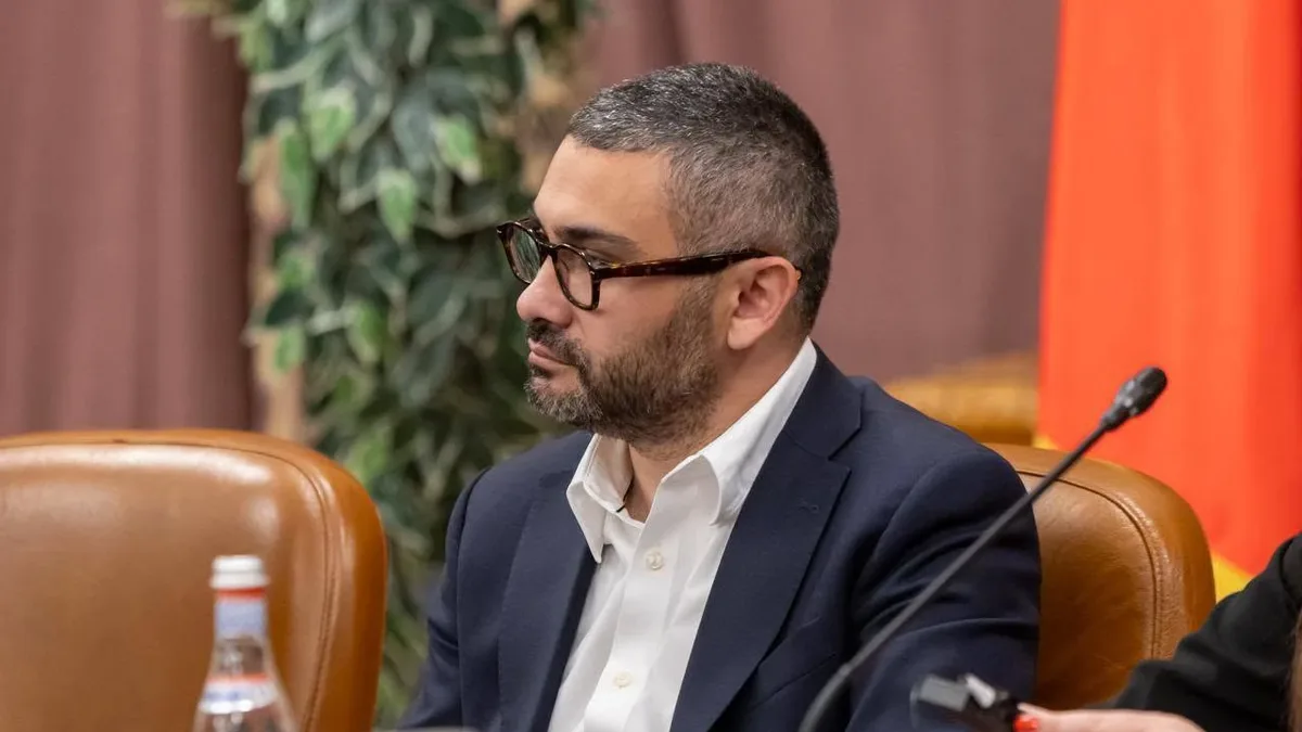 Florin Manole, cu ochelari și sacou închis la culoare, așezat la o masă lângă un microfon.
