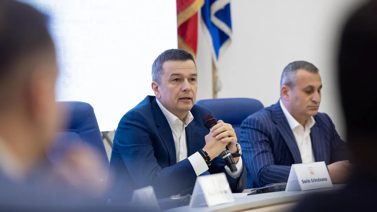 Sorin Grindeanu vorbește la un microfon ținut în mâini, așezat la masa unei reuniuni, lângă un alt participant.