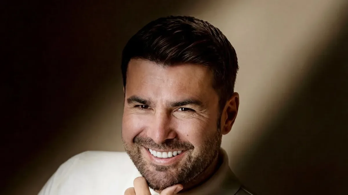 Adrian Mutu zâmbește, cu mâna sub bărbie, purtând un pulover crem pe un fundal estompat.