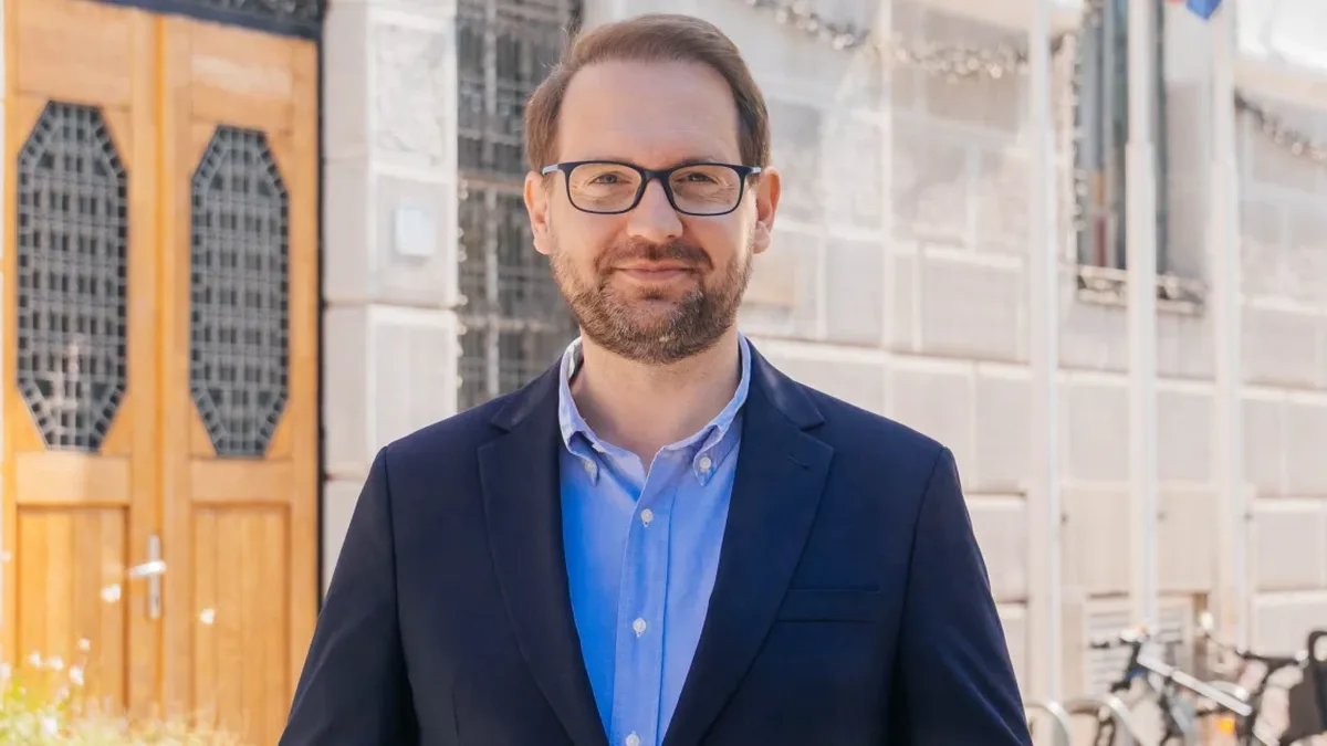 Dominic Fritz, cu ochelari și sacou bleumarin, în fața unei clădiri cu uși din lemn.