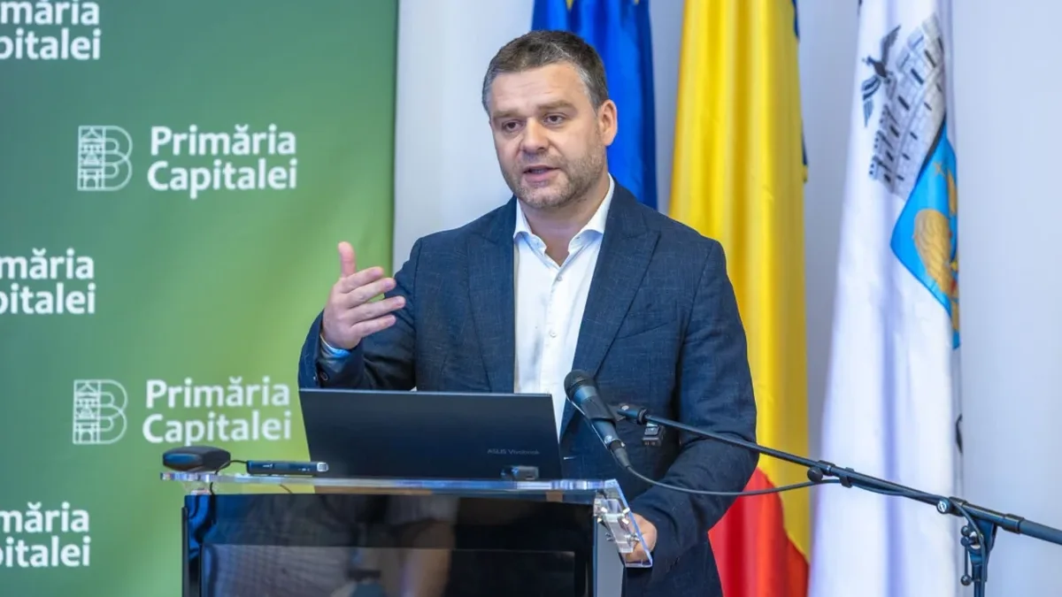 Ciprian Ciucu gesticulează la un pupitru cu laptop și microfon, în fața panoului Primăriei Capitalei și a drapelelor.