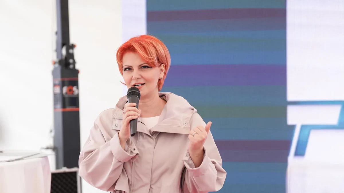 Lia Olguța Vasilescu vorbește la microfon și gesticulează, în fața unui ecran.