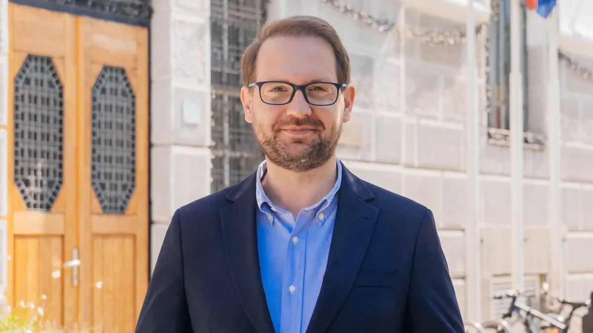 Dominic Fritz, cu ochelari, sacou bleumarin și cămașă albastră, în fața unei clădiri cu uși din lemn.