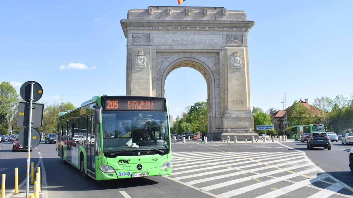 Autobuz verde STB în trafic, în fața Arcului de Triumf din București, sub un cer senin.