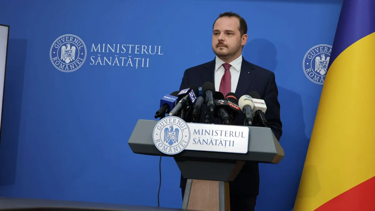 Alexandru Rogobete la pupitrul Ministerului Sănătății, în fața mai multor microfoane, lângă drapelul României.