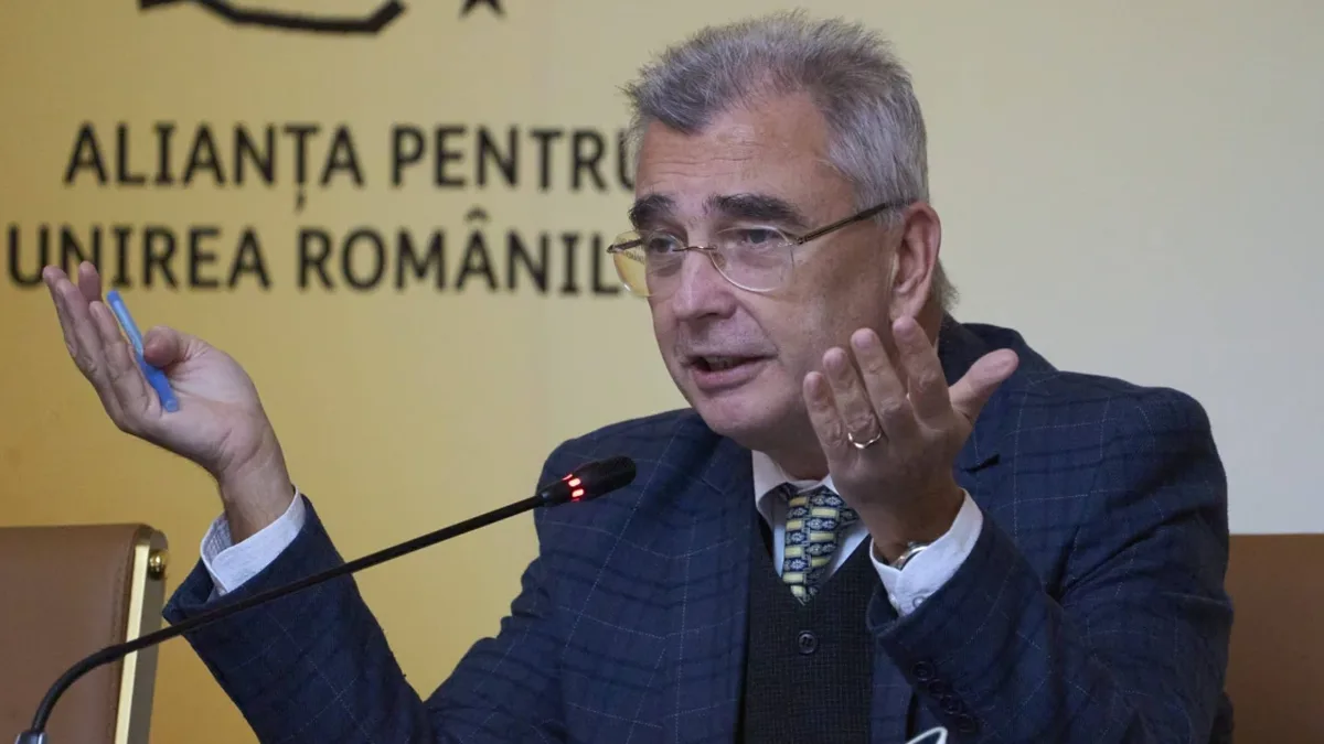 Petrișor Peiu gesticulează cu ambele mâini în fața unui microfon, pe fundalul unui panou al Alianței pentru Unirea Românilor.