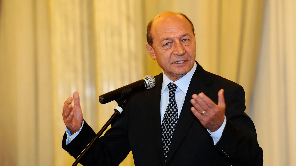 Traian Băsescu vorbește la microfon și gesticulează cu ambele mâini, în fața unor draperii deschise.