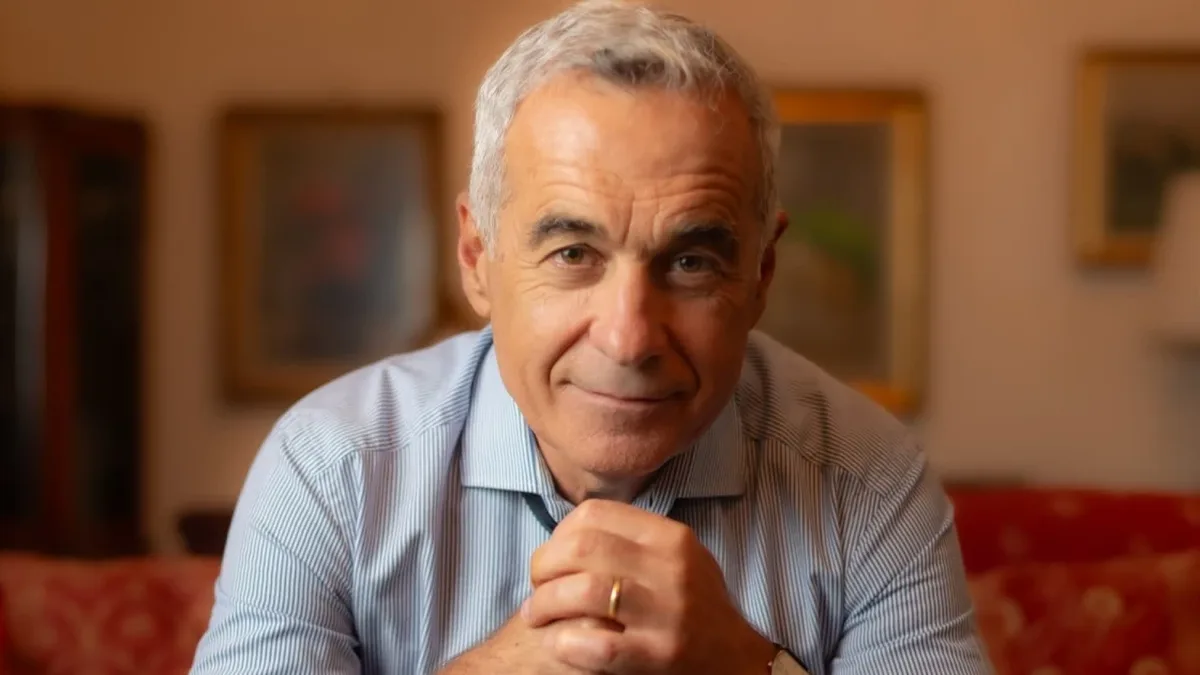 Călin Georgescu privește spre cameră, cu mâinile împreunate, într-un interior cu tablouri și o canapea roșie.