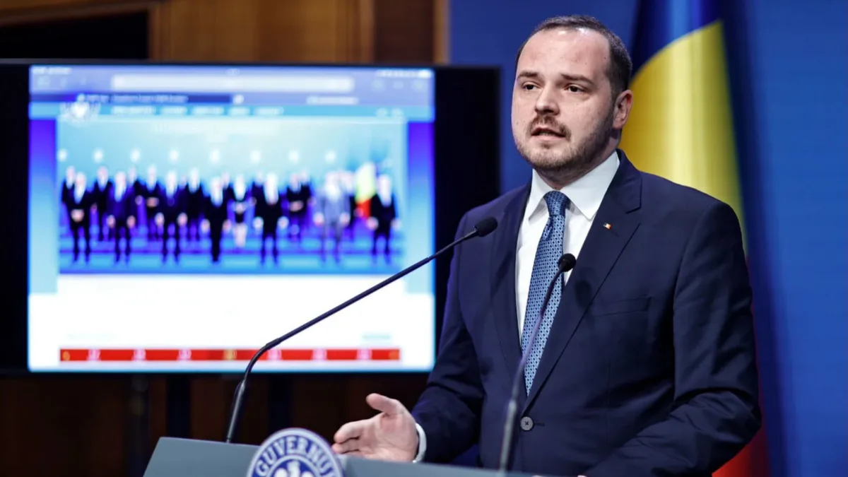 Alexandru Rogobete vorbește la un pupitru cu două microfoane, având în fundal un ecran și drapelul României.