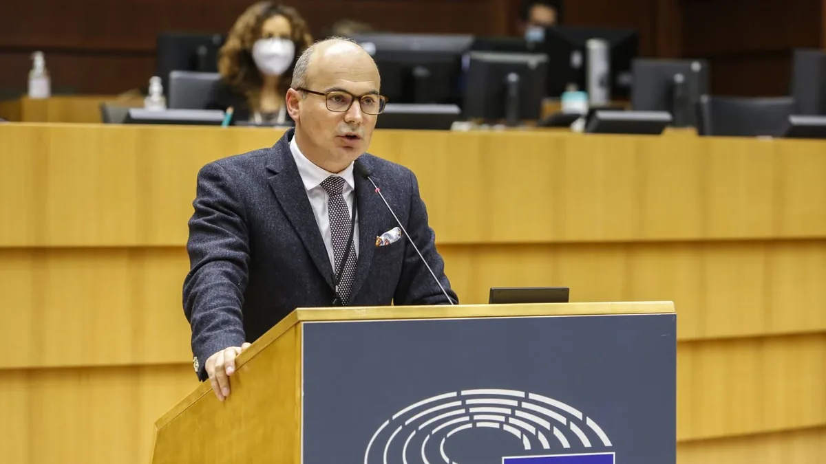 Rareș Bogdan vorbește la un pupitru cu emblema Parlamentului European, cu monitoare și participanți în fundal.