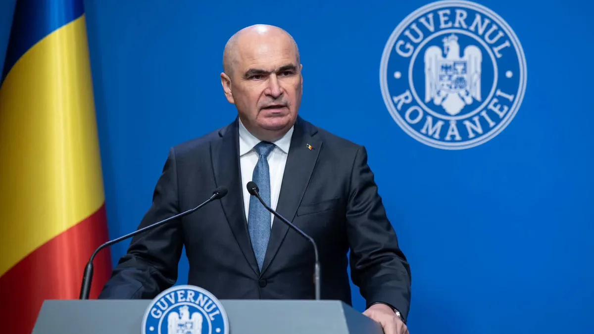 Ilie Bolojan vorbește la un pupitru cu două microfoane, lângă drapelul României, pe fundalul însemnelor Guvernului.