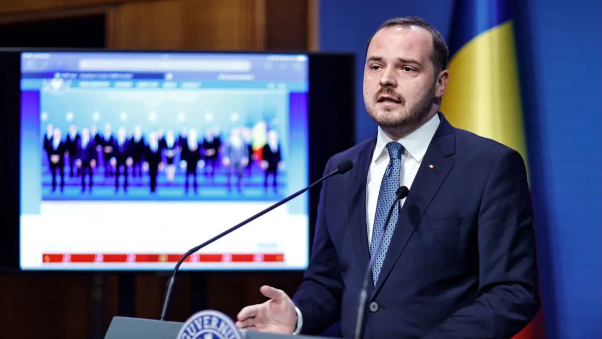 Alexandru Rogobete vorbește la un pupitru cu două microfoane, cu drapelul României și un ecran în fundal.