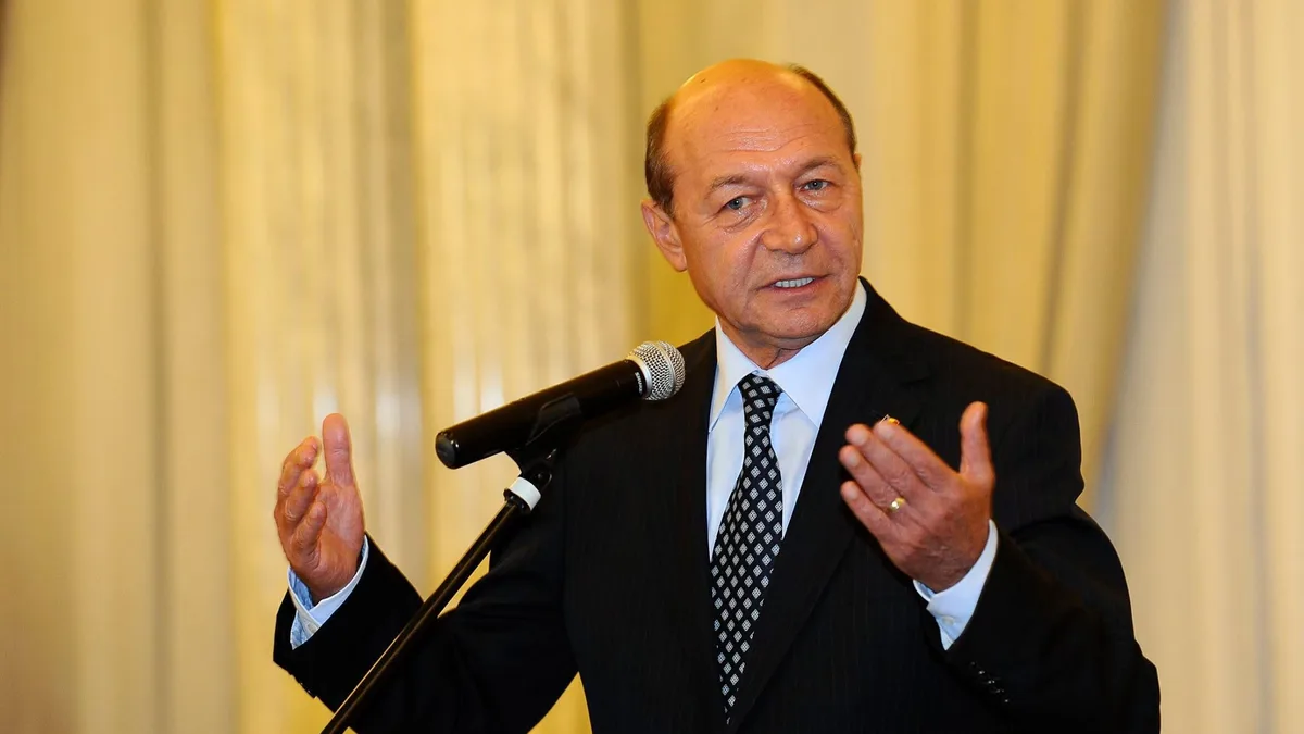 Traian Băsescu vorbește la microfon, gesticulând cu ambele mâini, în fața unor draperii deschise la culoare.