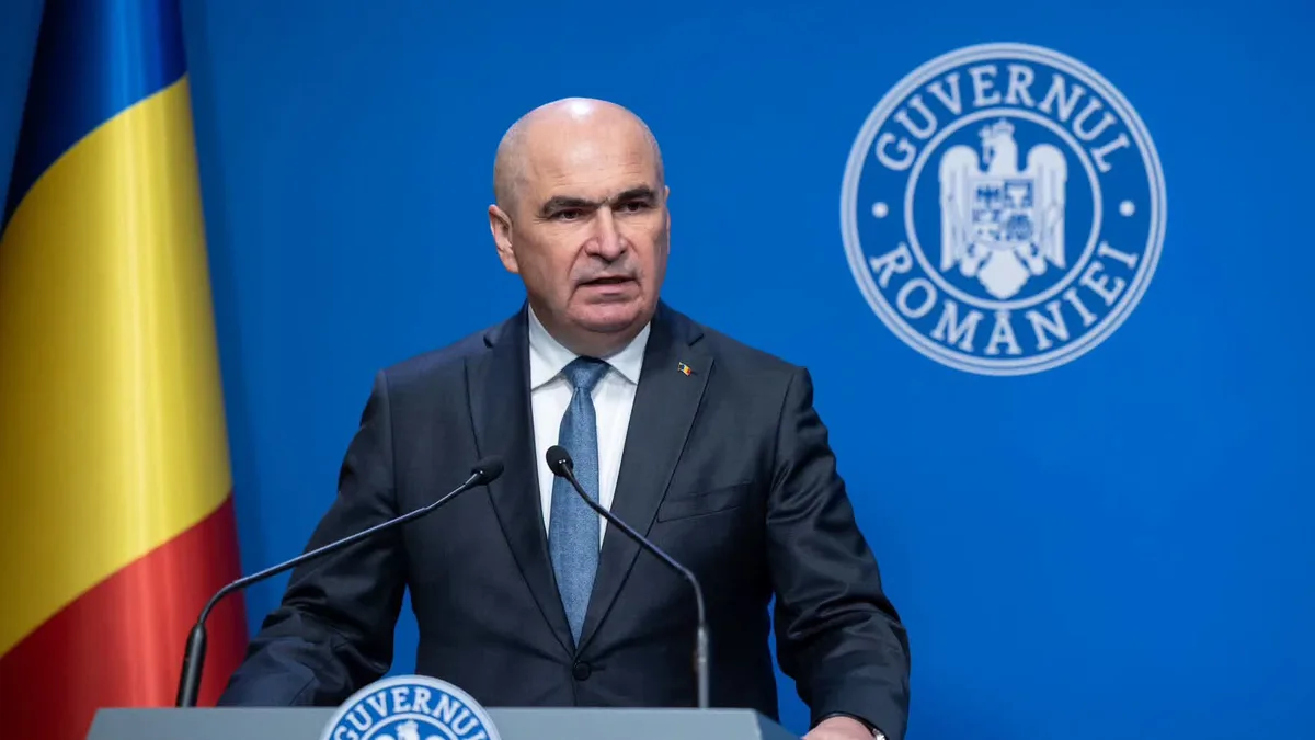 Ilie Bolojan la un pupitru cu două microfoane, lângă drapelul României, pe fundalul însemnelor Guvernului.