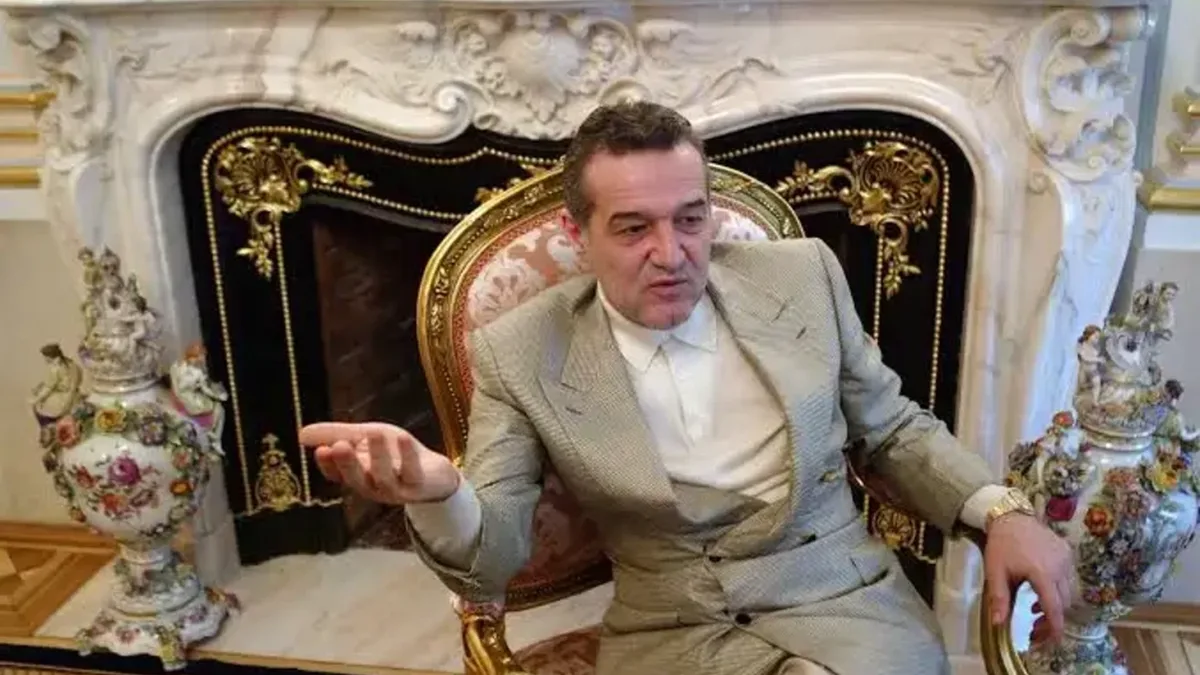 Gigi Becali, în costum deschis la culoare, gesticulează dintr-un fotoliu ornamentat, în fața unui șemineu.