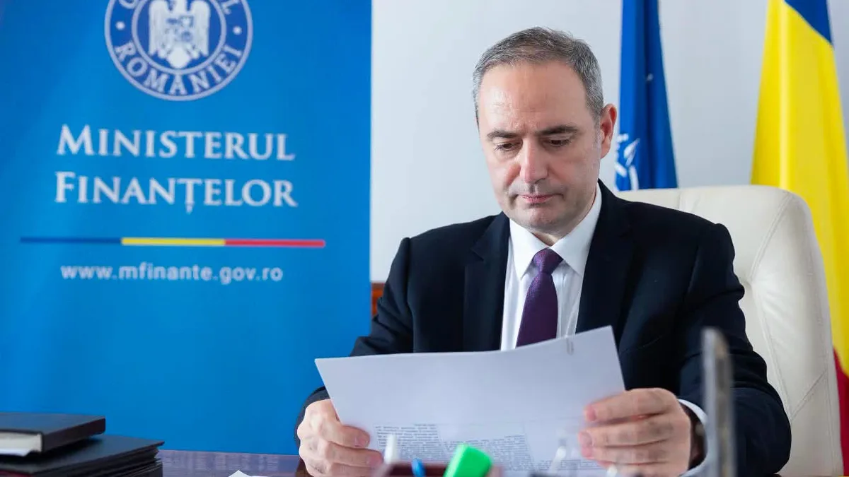 Alexandru Nazare consultă documente la birou, lângă un panou al Ministerului Finanțelor și drapelul României.