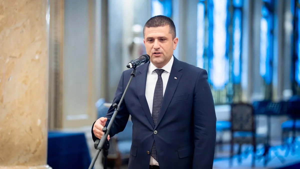 Radu Miruță vorbește la un microfon pe stativ și gesticulează într-o sală cu coloane și scaune albastre.