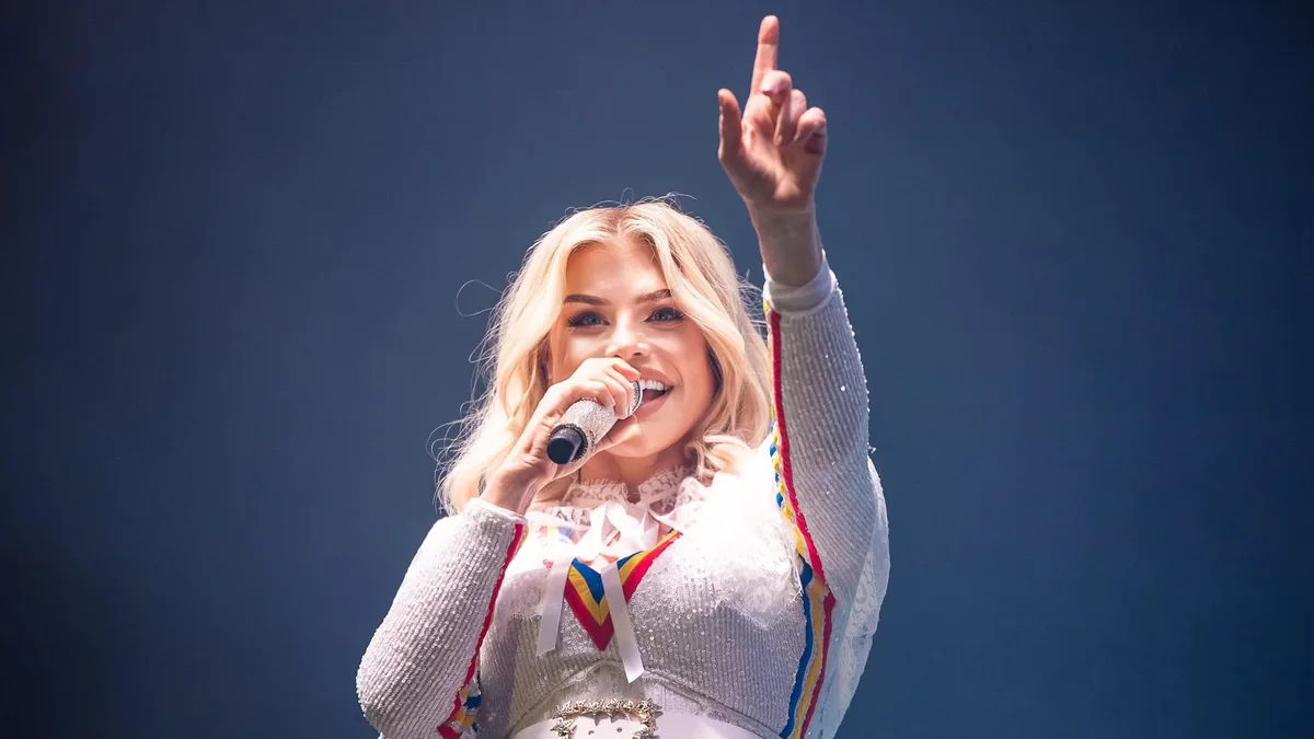 Loredana Groza cântă la microfon, cu un braț ridicat, într-o ținută albă cu detalii tricolore, pe fundal albastru.