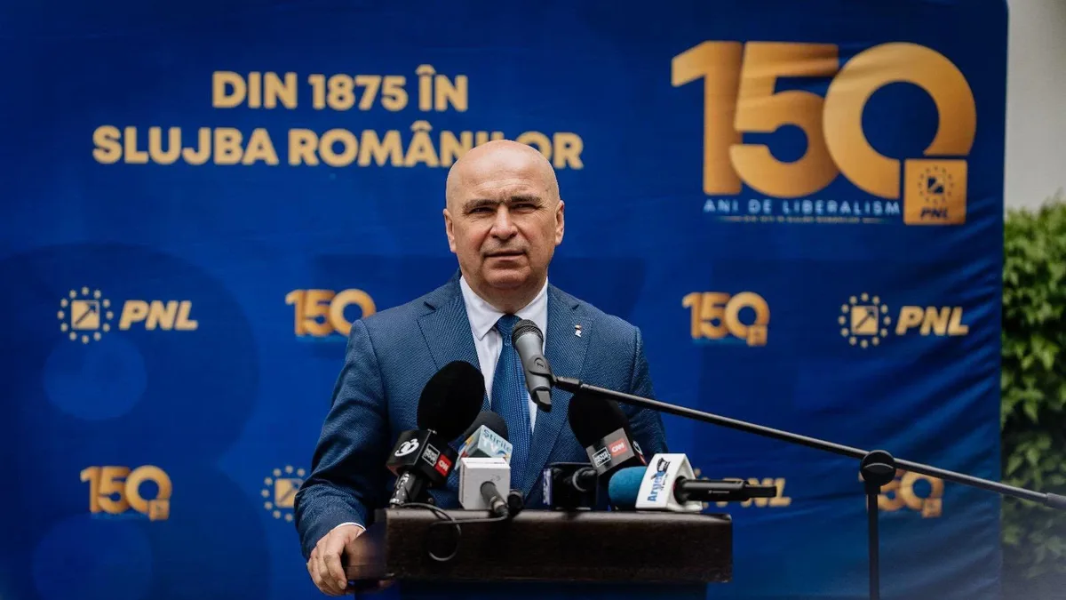 Ilie Bolojan la un pupitru cu microfoane, în fața unui panou PNL cu însemne aniversare.
