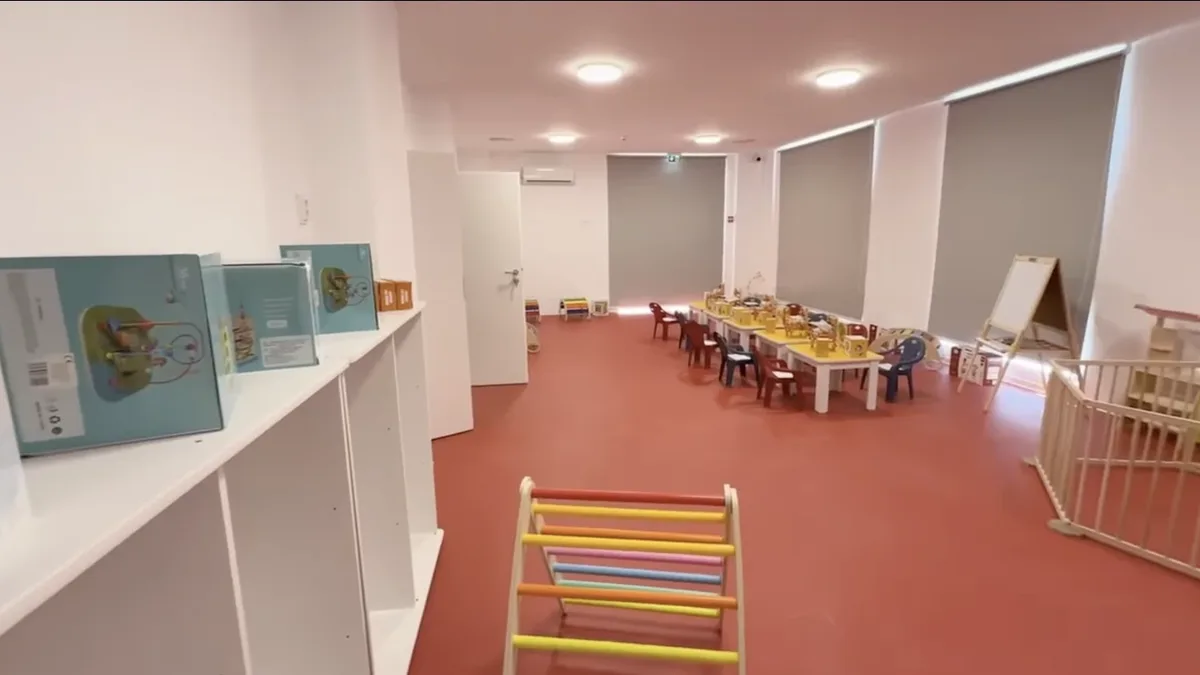 Sală fără persoane, cu mese și scaune pentru copii, rafturi cu materiale de joacă și un țarc din lemn.