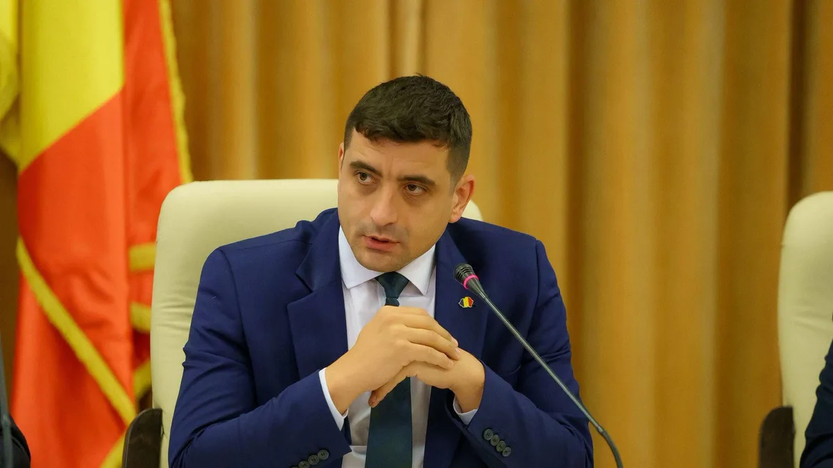 George Simion la masa unei reuniuni, cu mâinile împreunate lângă un microfon și un drapel românesc în fundal.
