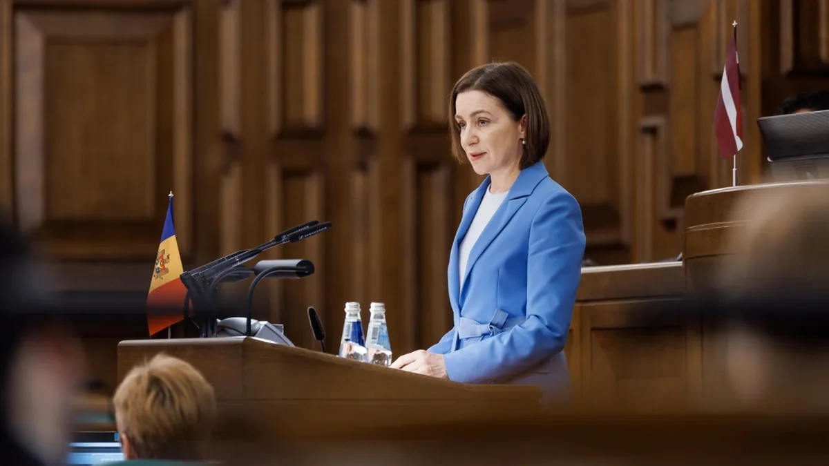 Maia Sandu vorbește la un pupitru cu microfoane, lângă drapelul Republicii Moldova, într-o sală cu lambriuri din lemn.