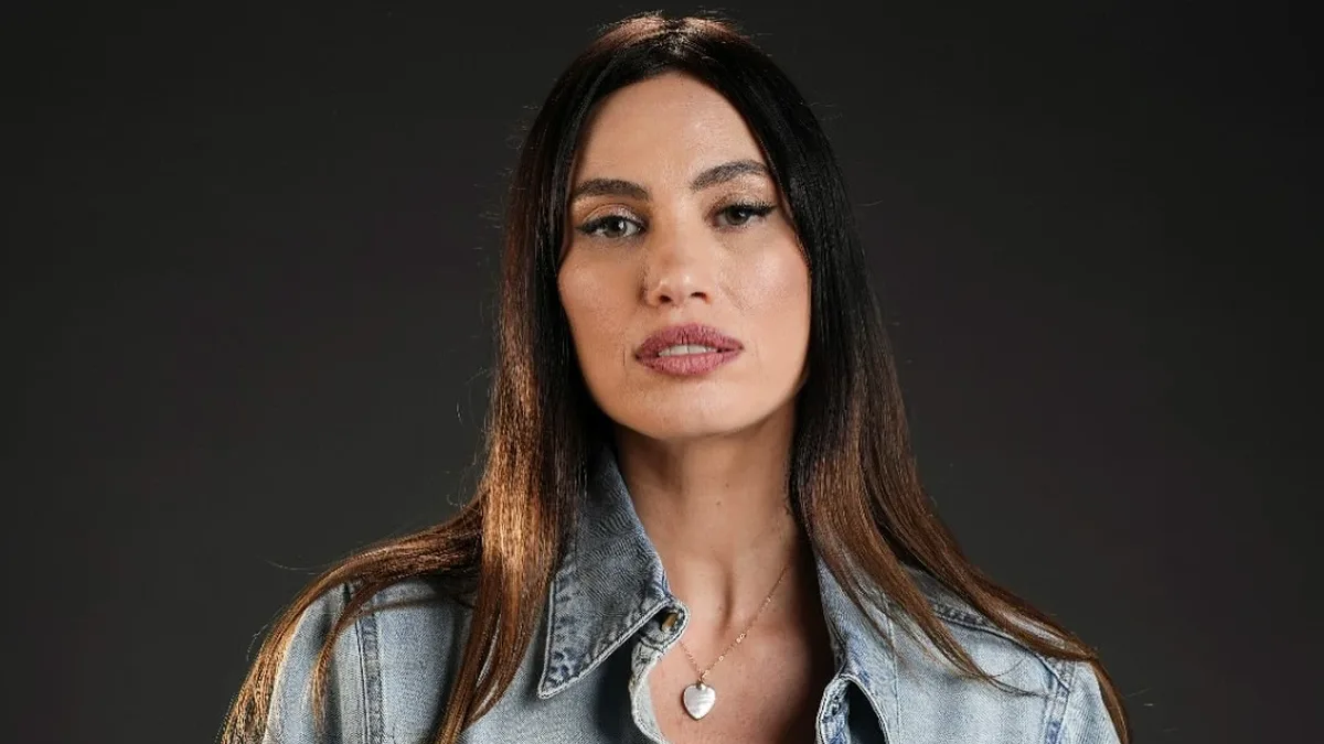 Alina Pușcău poartă o cămașă din denim legată în talie și privește spre cameră, pe un fundal închis.