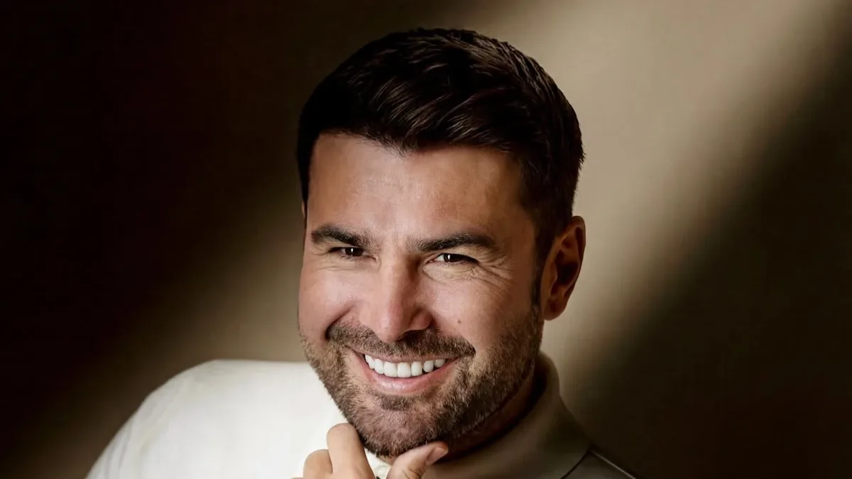 Adrian Mutu zâmbește spre cameră, purtând un pulover crem cu guler înalt și ținând mâna sub bărbie.