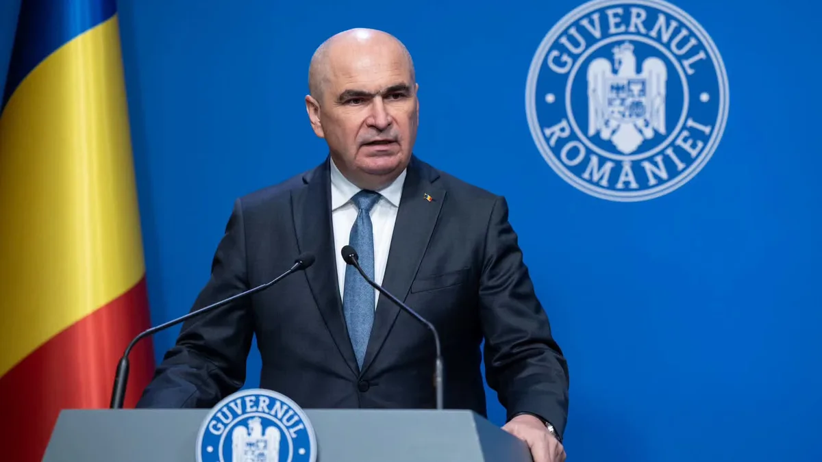 Ilie Bolojan vorbește la pupitru, lângă drapelul național, în fața însemnelor Guvernului României.