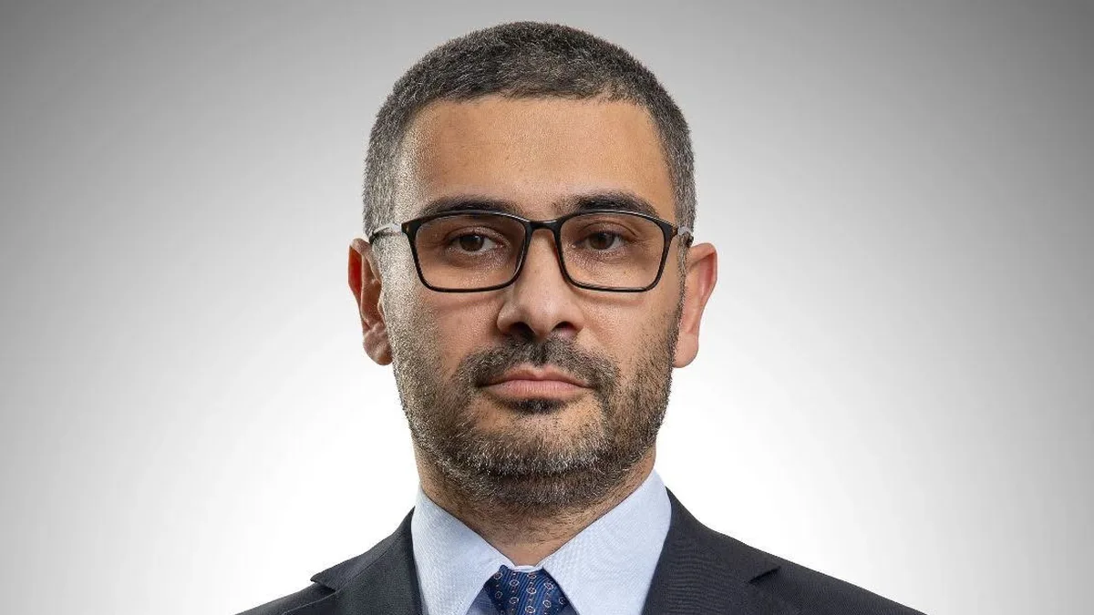 Florin Manole, cu ochelari, costum închis și cravată albastră, fotografiat frontal pe fundal gri.